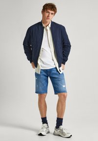 Cazadora bomber azul marino con bolsillos con cremallera, camiseta blanca, pantalones cortos de mezclilla con detalles desgastados, calcetines azul marino y zapatillas grises.