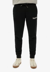 Zwarte sweatpants met een trekkoord in de taille, zijzakken en een wit "Superdry" logo op de linkerdij, met een zachte stoftextuur.