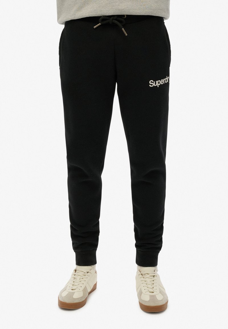 Zwarte sweatpants met een trekkoord in de taille, zijzakken en een wit "Superdry" logo op de linkerdij, met een zachte stoftextuur.