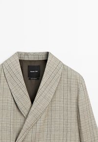 Blazer beige à carreaux avec col à revers et étiquette noire à l’intérieur indiquant Massimo Dutti, fabriqué au Portugal, sur fond blanc uni.