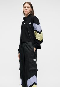 Femme portant un survêtement noir avec des rayures en chevron violet et jaune sur les manches et les jambes du pantalon, debout avec une main dans la poche.