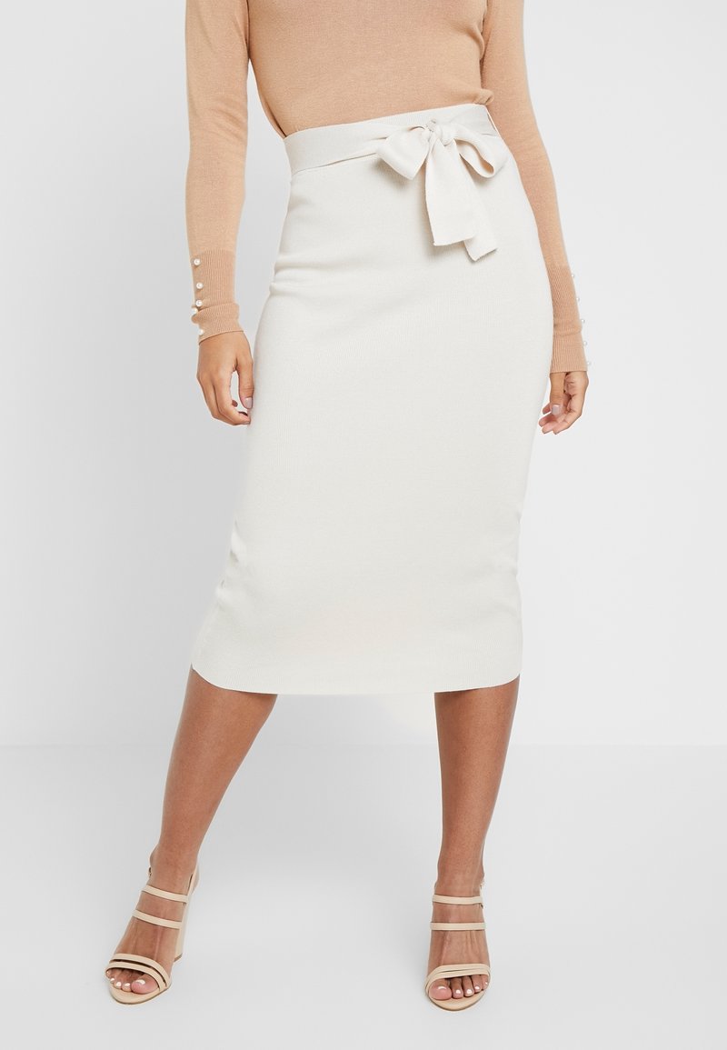 Femme portant une jupe blanche longueur genou avec un nœud noué à la taille, un haut beige à manches longues et des sandales à talons beiges à brides.