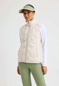Beige quiltad väst med hög krage och framskärp, bärs över en vit långärmad skjorta. Kompletteras av ljusgröna träningsleggings och en keps.