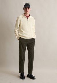 Jasno beżowy sweter z dzianiny z ćwierć zamkiem, oliwkowe spodnie z prążkowanej tkaniny, czarne skórzane buty i ciemna czapka. Prosty design i fakturowane materiały.