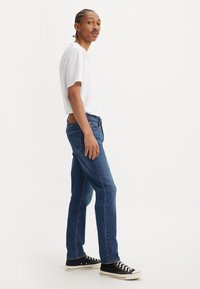 Mladý muž s vrkôčikmi, oblečený v bielom tričku, modrých džínsach a čiernych teniskách, stojí bokom na jednoduchom bielom pozadí.