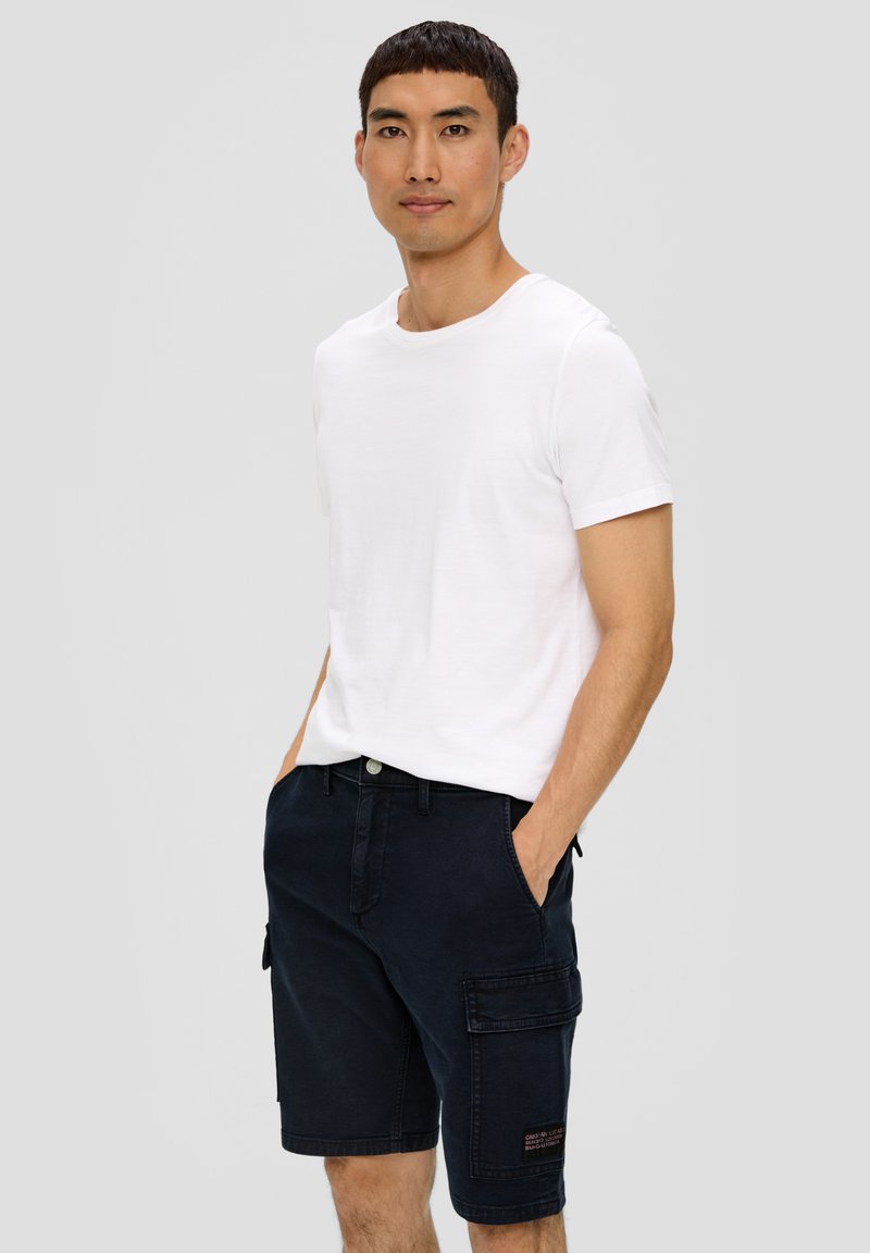 Biała koszulka z krótkim rękawem zestawiona z ciemnoniebieskimi szortami cargo, które mają boczne kieszenie i małą naszywkę z logo na lewej nodze.