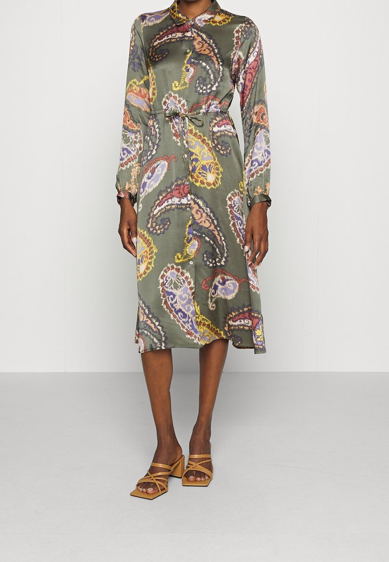 Femme portant une robe verte à manches longues boutonnée avec des motifs cachemire colorés et des sandales marron clair à talons blocs et brides.