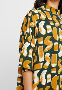 Zelené šaty s výraznými, abstraktními vzory v hořčicové a bílé barvě. Krátké rukávy, vysoký límec a měkká, strukturovaná látka.