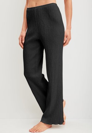 Personne pieds nus portant un pantalon noir texturé taille haute à jambes larges, debout devant un fond clair uni.