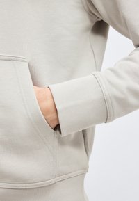 Main dans la poche avant d'un sweat à capuche beige avec poignet côtelé et détails de couture visibles sur un tissu doux.