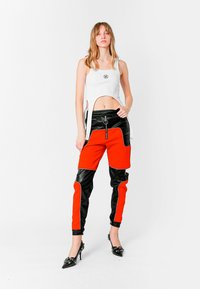 Mujer de pie con una camiseta corta blanca con lazos laterales, pantalones bicolores rojos y negros, y zapatos de tacón negro con hebillas decorativas.