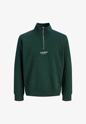 Ein blondes Junge steht nach vorne gerichtet und schaut nach links, trägt ein dunkelgrünes Sweatshirt mit Viertelreißverschluss, braune lockere Hose und beige Turnschuhe.