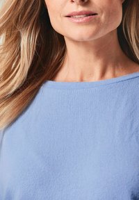 Himmelblaues, strukturiertes Stoffhemd mit weitem Ausschnitt, gesehen vor einem teilweise sichtbaren Hals und fließendem Haar.