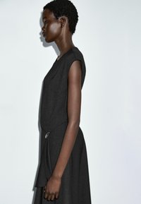 Schwarzes kurzärmeliges Kleid mit lockerer Passform, das an der Seite eine Drapierung und einen runden silbernen Akzent in der Taille hat. Flacher Hintergrund.