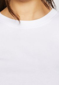 Gros plan d'une personne portant un t-shirt blanc uni à col rond avec des cheveux bruns partiellement visibles.