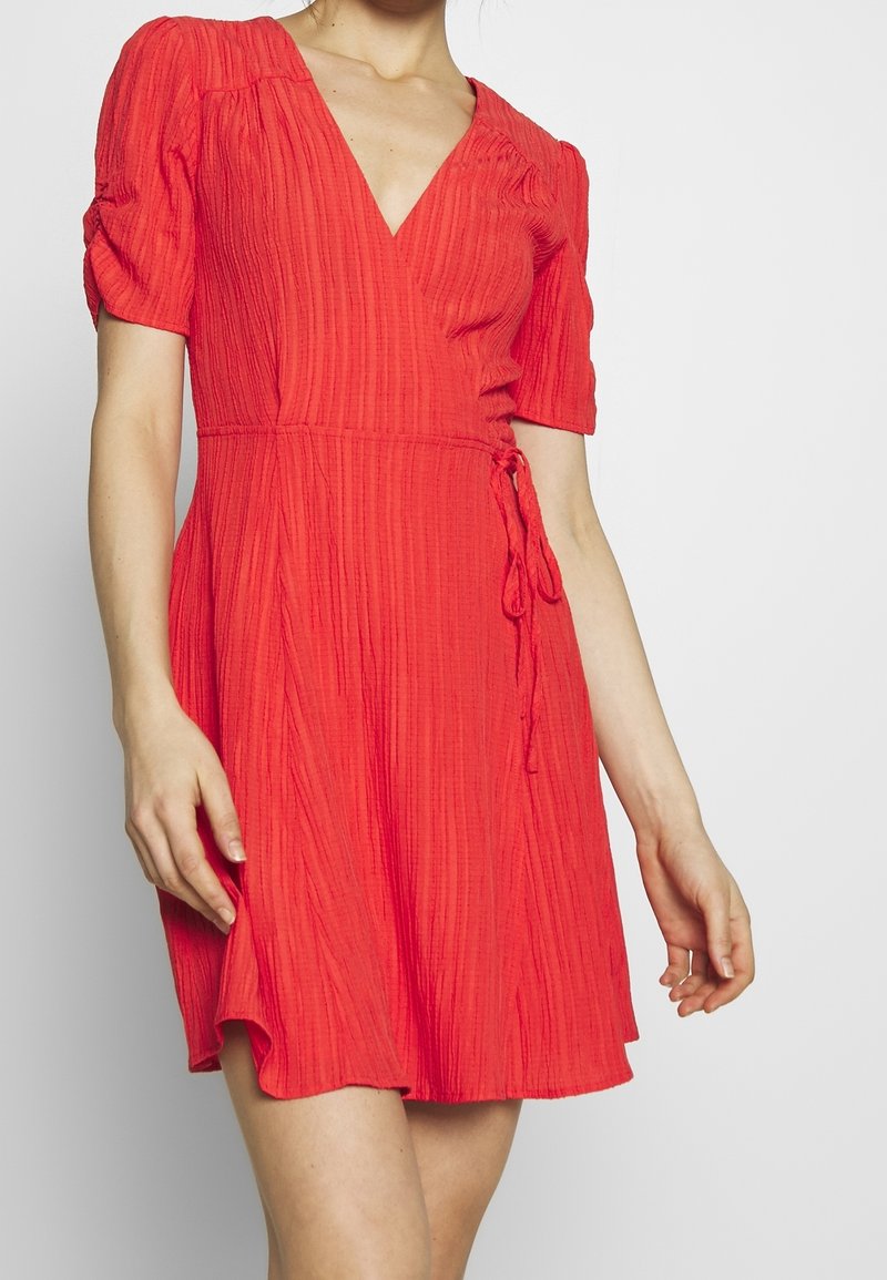 Femme portant une robe portefeuille rouge texturée avec des manches courtes bouffantes et une ceinture à la taille, debout devant un fond clair uni.