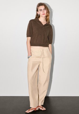 Polo en tricot marron à manches courtes, pantalon beige à cordon de serrage et sandales plates marron et rouge. Tissu doux et coupe décontractée.