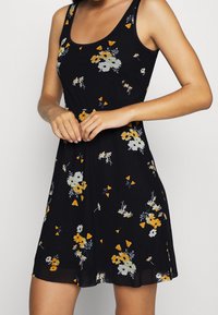 Vestido floral de tela negra con flores amarillas y blancas, un torso ajustado y una falda acampanada. Diseño sin mangas con escote redondo.