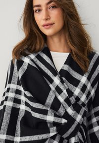 Sort og hvid plaid sjal lavet af blødt stof, med et stort ternet mønster og et draperet design, båret omkring skuldrene.