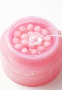 Lyserød krukke med Medicube kollagenkapselcreme, fyldt med små, runde lyserøde kapsler. En gennemsigtig ske holder en kapsel over krukken.