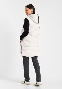 Lichte beige puffer vest met een capuchon, zijzakken en een gequiltte textuur. Combinatie met donkere jeans en witte sneakers.