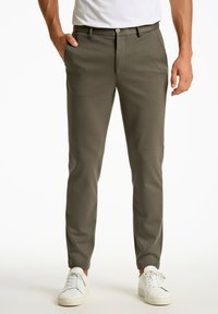Chinos kaki fabriqués dans un tissu structuré, avec une coupe slim, des poches latérales et une fermeture à bouton. Portés avec des baskets blanches.