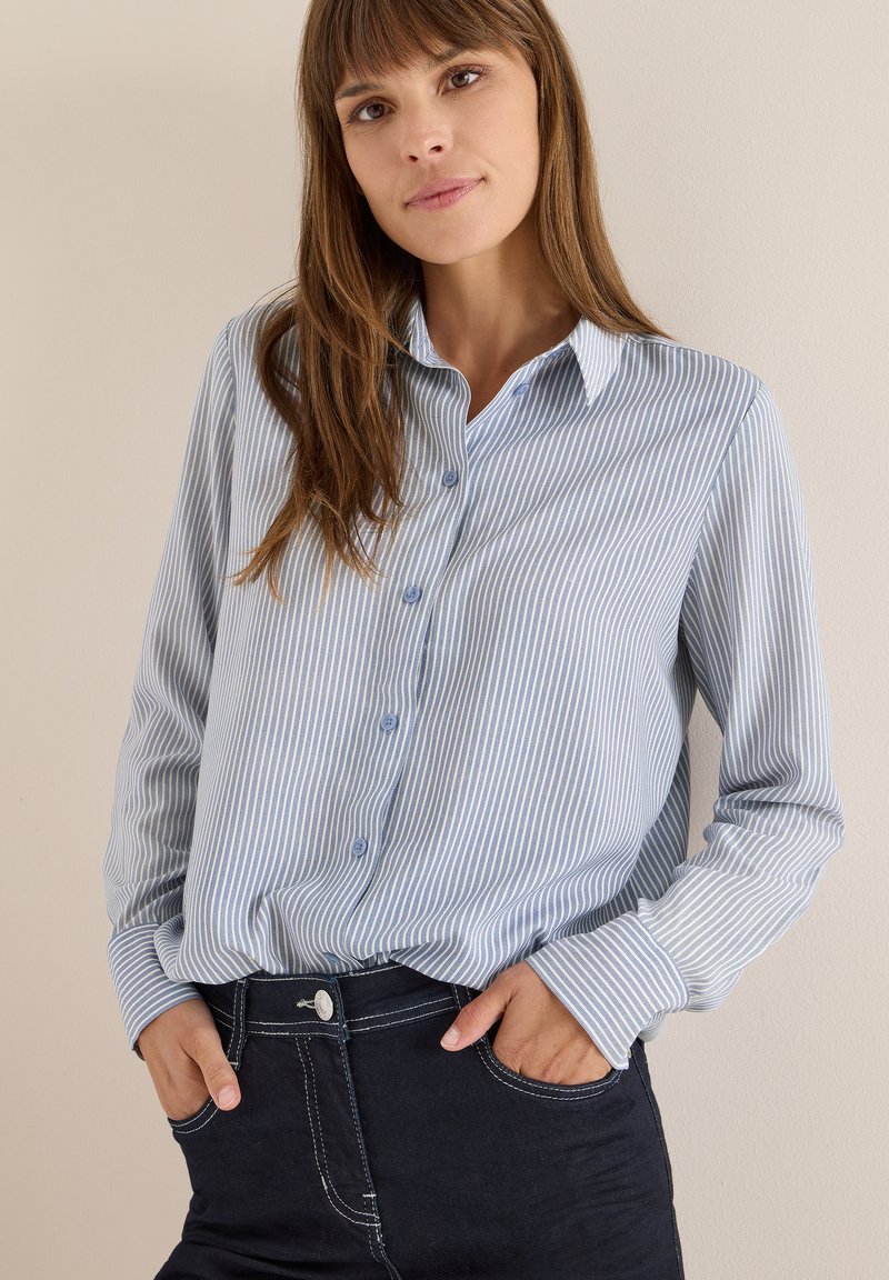 Camicia button-up a righe blu chiaro con maniche lunghe, caratterizzata da un colletto e bottoni blu, abbinata a jeans scuri e un look casual.