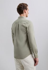 Uomo che indossa una camicia lunga verde chiaro con bottoni e pantaloni grigio chiaro, rivolto di spalle su uno sfondo grigio uniforme.