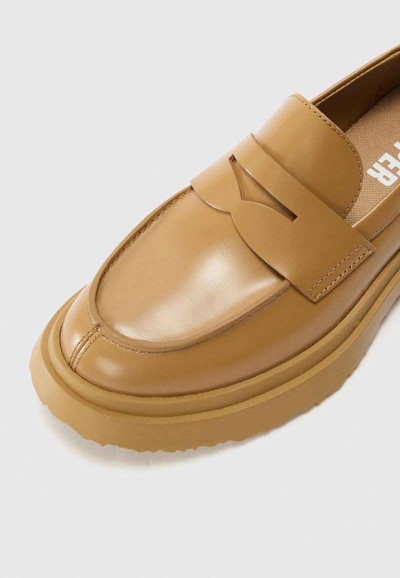 Mocasín de cuero marrón con punta redonda, que cuenta con una tira decorativa y una suela de goma gruesa y texturizada. Acabado liso y detalles de costura sutiles.