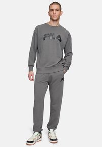 Grijze katoenen sweatshirt met zwart FILA-logo, gecombineerd met bijpassende grijze sweatpants met een logo. Model draagt multikolor sneakers.