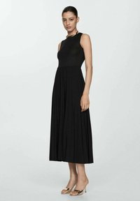 Schwarzes, ärmelloses Maxikleid mit tailliertem Oberteil und plissiertem Rock. Glattes, strukturiertes Material. Das Model trägt metallische offene High Heels.