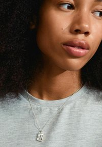 Sølvvedhængsnor på en fin kæde, båret over en lys grå t-shirt. Nærbillede der viser detaljerne på vedhænget og nakkeområdet.