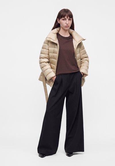 Femme portant une doudoune beige sur une chemise marron avec un pantalon noir à jambes larges et des chaussures noires, debout les mains dans les poches.