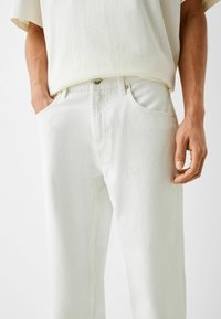 Pantalones de denim blancos con corte recto, que cuentan con bolsillos frontales y un cierre de botón metálico en la cintura, tejido texturizado en toda su superficie.
