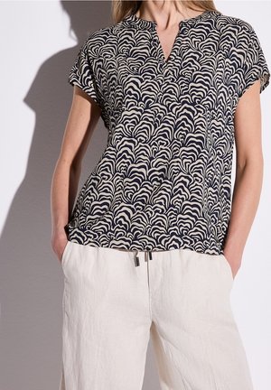 Vrouw draagt een zwart-wit blouse met abstract patroon en korte mouwen, met witte, loszittende broek, handen in de zakken, tegen een effen achtergrond.