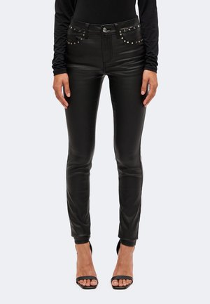 Mannequin portant un pantalon noir ajusté effet cuir avec des poches cloutées, un haut noir et des chaussures à talons ouvertes à l’avant.