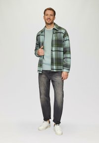 Grün und blau karierte Hemd, hellblauer Pullover, graue Jeans und weiße Sneakers. Das Model steht mit einer Hand in der Tasche.