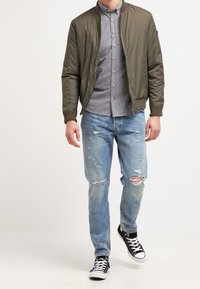 Giacca bomber verde oliva sopra una camicia a quadri grigia, abbinata a jeans chiari strappati e sneakers nere a basso profilo.