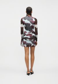 Άτομο με πλεξούδες, που φορά κοντό φόρεμα με μακριά μανίκια και λουλουδένιο μοτίβο, δεμένο στη μέση, και μαύρες παπούτσια με χαμηλό τακούνι, στέκεται σε απλό φόντο.