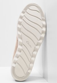 Semelle d'une chaussure beige avec des motifs de semelle ondulés et le logo "Clarks" embossé au centre, reposant contre un fond blanc.