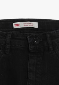 Vaqueros de mezclilla negra con cintura alta, ajuste super ajustado, cierre de botón visible y una etiqueta blanca con texto rojo para la identificación de la marca.