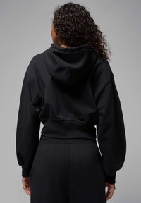 Mujer con cabello rizado vistiendo una sudadera corta negra y pantalones negros a juego, de espaldas frente a un fondo gris liso.