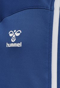 Blå atletisk jakke i tekstureret stof med en hvid lynlås og logo. Logoet viser et stiliseret bi-design med "hummel" tekst nedenfor.