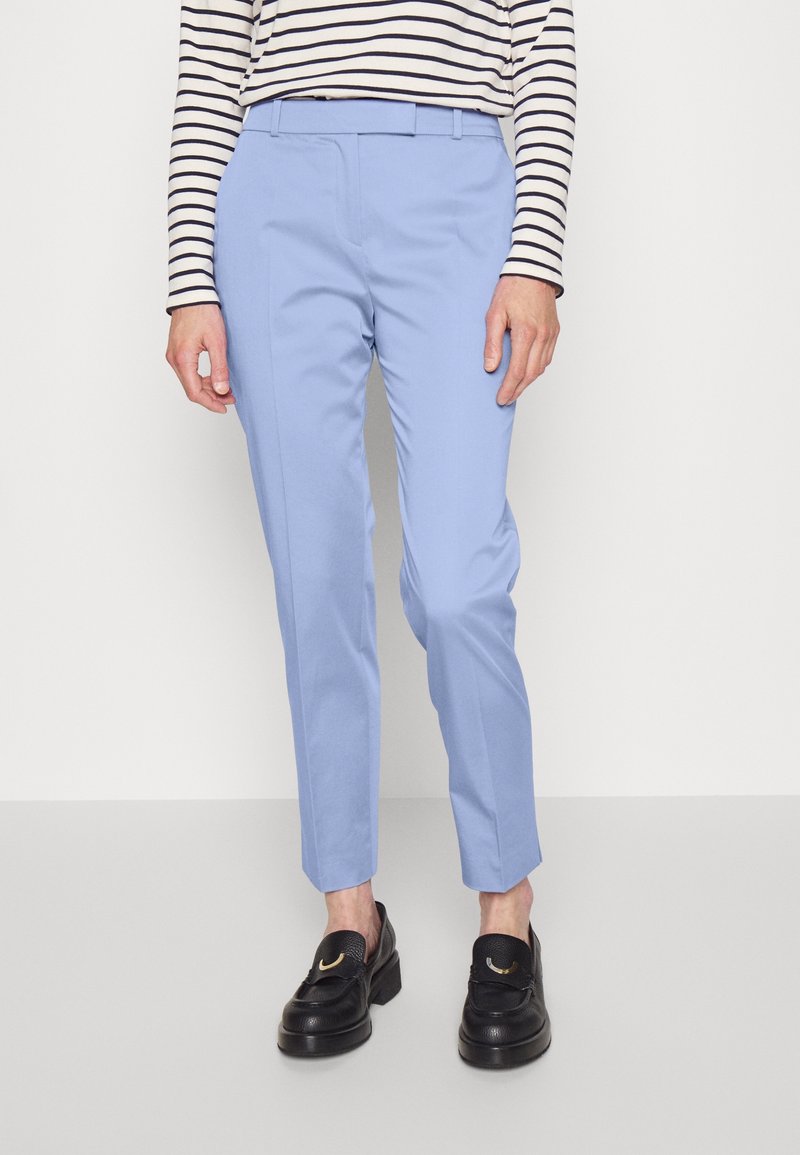 Hellblaue, taillierte Hose mit schmaler Passform, aus glattem Stoff gefertigt, mit Gürtelschlaufen und vorn angewendeten Bügelfalten. Kombiniert mit schwarzen Slippern.