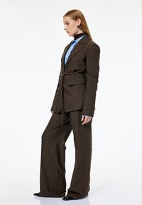 Brunternetternet skræddersyet jakkesæt med bælte i taljen, der har en blå skjorte under. Slanke bukser og spidse sorte sko.