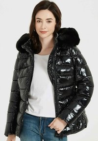 Doudoune noire avec finition brillante, capuche en fausse fourrure, design matelassé, fermeture éclair à l'avant et fermeture à pression. Portée par-dessus un haut blanc.