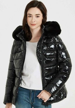 Doudoune noire avec finition brillante, capuche en fausse fourrure, design matelassé, fermeture éclair à l'avant et fermeture à pression. Portée par-dessus un haut blanc.