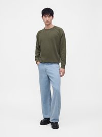 Jeune homme aux cheveux noirs porte une chemise verte à manches longues, un jean large bleu clair et des chaussures noires, debout devant un fond blanc.