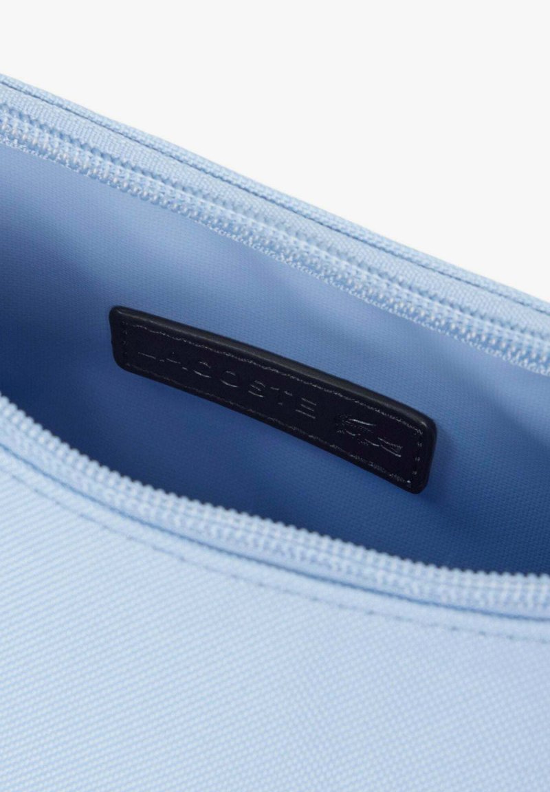 Petite pochette bleu clair zippée ouverte pour montrer l'étiquette de marque Lacoste bleu foncé à l'intérieur près de la fermeture éclair.