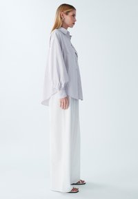 Gestreiftes, helllila Übergröße-Hemd mit Kragen, durchgehender Knopfleiste und langen Glockenärmeln, kombiniert mit weißen weit geschnittenen Hosen und Sandalen.